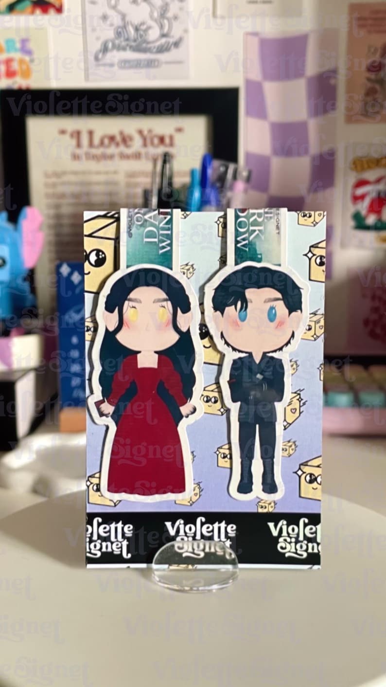 May include: A bookmark featuring two cartoon figures, a woman in a red dress and a man in a black suit. The bookmark has a light blue background with a pattern of small cheese slices. The text "Violette Signet" is visible.