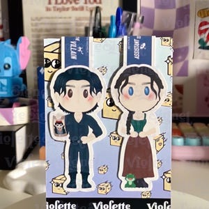May include: A decorative card featuring two cartoon characters, one in a dark outfit and the other in a green and brown dress. The card has a light blue background with small illustrations and the text "VIOLETTE SIGNET".