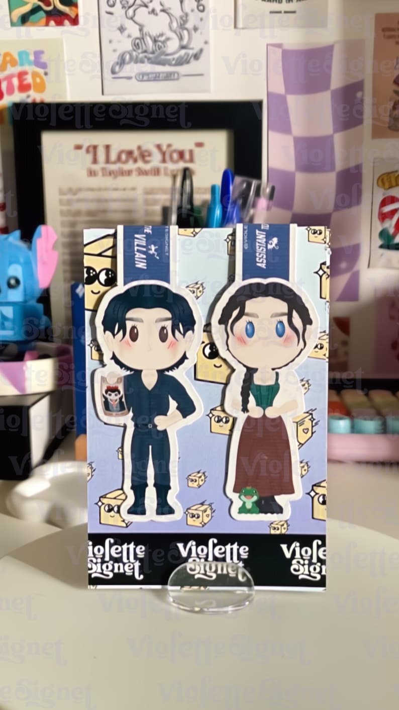 May include: A decorative card featuring two cartoon characters, one in a dark outfit and the other in a green and brown dress. The card has a light blue background with small illustrations and the text "VIOLETTE SIGNET".