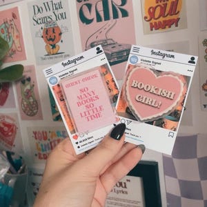 Puede incluir: Dos pegatinas estilo Instagram. Una presenta un corazón rosa con el texto "BOOKISH GIRL!". La otra muestra un bloc de notas rosa con el texto "SO MANY BOOKS SO LITTLE TIME". El fondo incluye varias obras de arte mural.