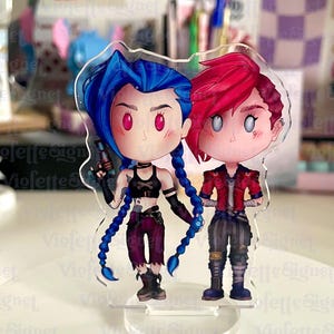 Puede incluir: Soporte acrílico transparente con dos personajes de dibujos animados con cabello azul y rojo, tomados de la mano. Los personajes visten atuendos detallados, uno con top negro y pantalón morado, el otro con chaqueta roja y pantalón a rayas. El soporte está sobre una superficie blanca.