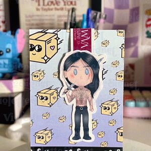 May include: A sticker featuring a cartoon illustration of a character holding a knife, with a partially exposed torso and black pants. The background has repeating images of smiling boxes. The sticker is displayed on a clear stand.