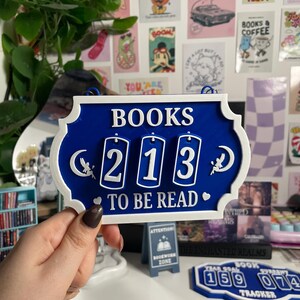 May include: A blue and white sign with the words "BOOKS TO BE READ" and the number "213". The sign has a decorative border and small angel and moon designs. A smaller sign reads "BOOKWORM ZONE".