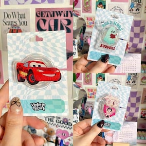Puede incluir: Una colección de agarres de teléfono de acrílico transparente con diseños de dibujos animados. Un agarre muestra un coche de carreras rojo, otro un fantasma leyendo un libro con el texto "Read More" y "BOOOOKS", y un tercero presenta un personaje de dibujos animados.