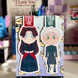 May include: Two illustrated character bookmarks. One features a woman in a dark dress with a red bow, the other a man in a dark suit with a white cat. The bookmarks are on a light blue background with the text "Viofette Signet".