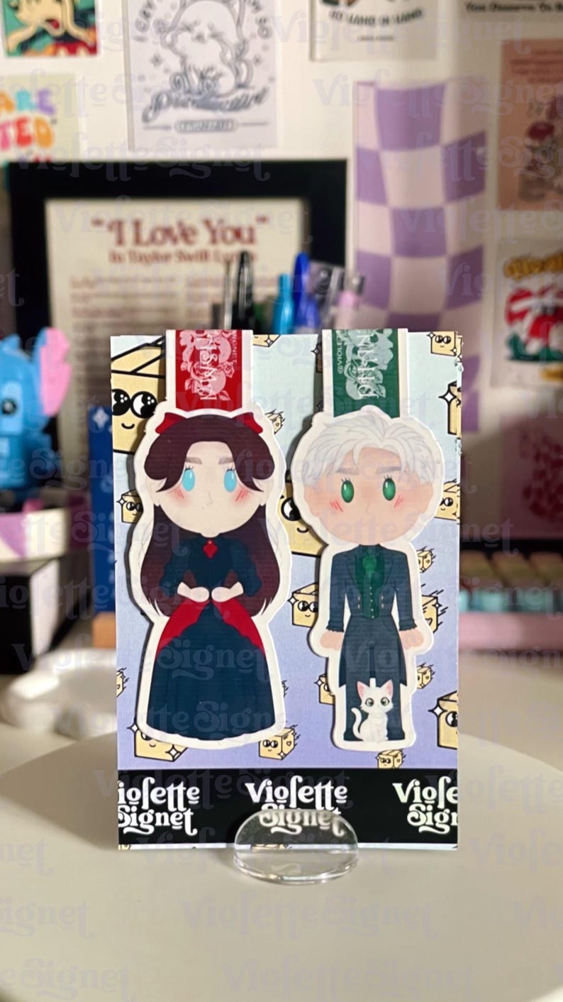 May include: Two illustrated character bookmarks. One features a woman in a dark dress with a red bow, the other a man in a dark suit with a white cat. The bookmarks are on a light blue background with the text "Viofette Signet".