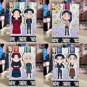 May include: Four character-themed bookmarks with cartoon illustrations. Each bookmark features a different pair of characters, with a purple background and the text "Violette Signet". The bookmarks are displayed on clear stands.