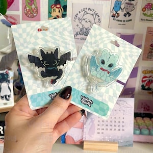 Puede incluir: Dos pines acrílicos transparentes con dibujos de dragones. Uno es negro con detalles rojos, el otro es azul claro con una bufanda roja. Cada pin está en una tarjeta con un patrón de cuadros y el texto "Violette Signet".