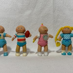 May include: Four Cabbage Patch Kids dolls, each with a different outfit and accessories. The dolls are all wearing different colored clothing, including blue, pink, and white. The dolls are all smiling and have blonde hair.
