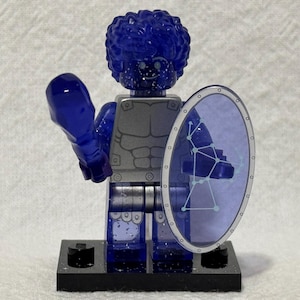 May include: A blue and silver Lego minifigure with a blue shield and a blue weapon. The minifigure has a blue head with a silver face and a silver torso with a blue pattern. The shield has a blue and white pattern.