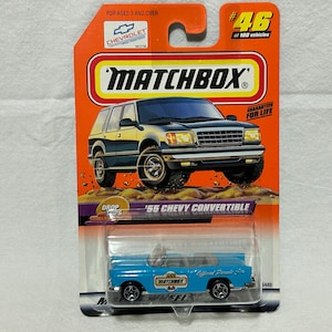 May include: A blue 1955 Chevrolet convertible toy car in a Matchbox die-cast car package. The car has a white roof and a red and white stripe on the side. The package is orange and has a picture of a Chevrolet SUV on the front. The package also has the text "Drop Tops" and "'55 Chevy Convertible".