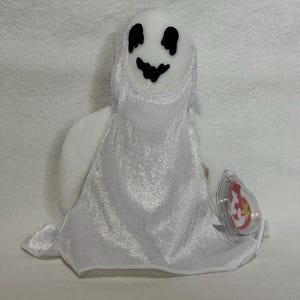May include: White plush ghost with a black smiling face. The ghost is wearing a white satin gown and has a red and white Ty Beanie Baby tag attached.
