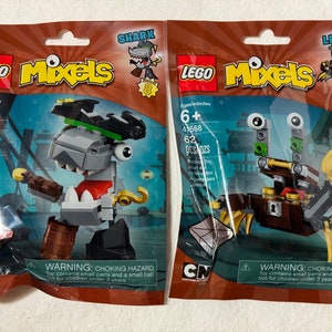 May include: Two unopened LEGO Mixels bags, one featuring a grey shark character named Sharx and the other a brown crab character named Lewt. Both bags have the LEGO logo, the Mixels logo, and the Cartoon Network logo.