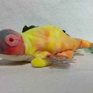 May include: A colorful tie-dye chameleon plush toy with a red eye and a green leafy crest. The toy has a yellow, orange, pink, and blue tie-dye pattern.