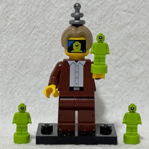 May include: A brown Lego minifigure with a yellow face and a black antenna on its head. The minifigure is holding a green Lego minifigure with a yellow face. There are two other green Lego minifigures on the base.