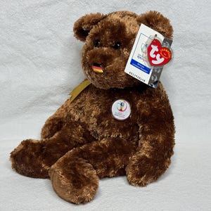 May include: A brown teddy bear with a German flag on its nose and a FIFA World Cup emblem. The bear has a Ty tag attached. The bear is sitting on a white surface.
