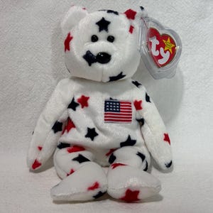 May include: White plush teddy bear with red and blue stars and an American flag patch. The bear has a red heart-shaped tag with the words "Ty Inc." and "Beanie Babies" on it.