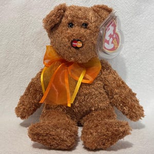 May include: A brown teddy bear with a bright orange ribbon around its neck. The bear has a red heart-shaped tag on its chest with the word "Ty" on it. The bear is wearing a clear plastic tag with the Ty logo and the words "Beanie Babies" on it.