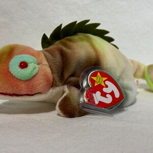 May include: A green, brown, and white camouflage patterned Ty Beanie Baby chameleon plush toy with a red eye and a green eye. The toy has a Ty Beanie Baby tag attached to it.