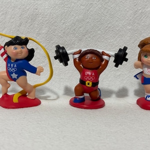 May include: Three vintage figurines of athletes in Olympic uniforms. One is holding a jump rope, another is lifting weights, and the third is holding a baton. Each figurine is on a red base and wears a USA uniform.