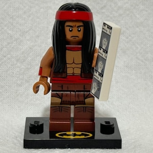 May include: Lego minifigure of a Native American man with long black hair, wearing a red headband, a brown vest, and brown pants. He is holding a white rectangular object with black images on it. The minifigure is standing on a black base with a yellow Batman logo.