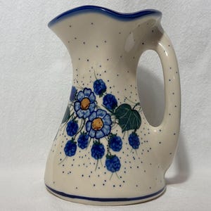 May include: A white ceramic pitcher with a blue and white floral design. The pitcher has a wide mouth and a handle. The floral design features blue flowers with yellow centers and blue berries.