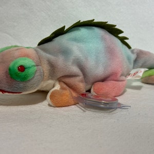 May include: A tie-dye green, blue, and pink chameleon plush toy with a red eye. The toy has a green and white tag attached to it.
