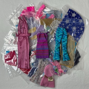 May include: A selection of doll clothes in clear plastic bags. Items include a pink dress, a purple and blue patterned dress, blue and green patterned trousers, and a blue star-patterned cape.