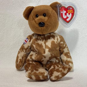 May include: A brown and tan camouflage patterned beanie baby bear with a red, white and blue Union Jack patch on its arm. The bear has a red Ty Beanie Babies tag attached to its ear.