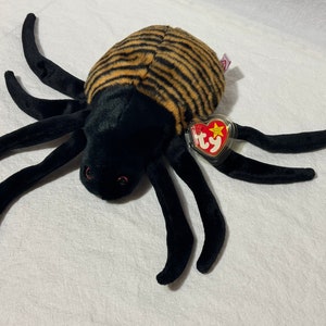 May include: A plush spider toy with a black body and eight long, black legs. The spider's back is brown with black stripes. It has small red eyes and a TY heart-shaped tag.