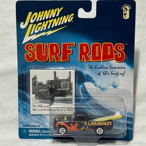 May include: A black and orange toy pickup truck with a surfboard on the back. The truck has the words "Lava Mama's" printed on the side. The truck is on a blue and white background with the words "Surf Rods" and "The Endless Summers of '60s Surfing!" printed on it.