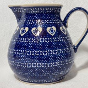 May include: A blue and white ceramic pitcher with a floral pattern and a heart design. The pitcher has a wide mouth and a handle.
