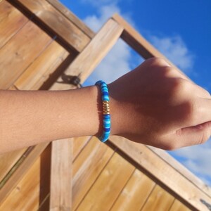 May include: A blue and gold beaded bracelet with a gold accent. The bracelet is on a person's wrist.
