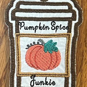 May include: Embroidered coffee cup ornament with a pumpkin spice design. The cup has a brown rim and a white background with the text "Pumpkin Spice" and "Junkie".