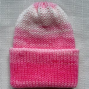 Può includere: Un berretto lavorato a maglia in un gradiente di rosa e bianco. Il cappello presenta un bordo ripiegato e un aspetto morbido e strutturato. Il colore passa da un rosa brillante sul bordo al bianco sulla parte superiore.