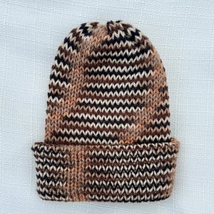 May include: A knitted beanie hat with a brown, black, and cream zigzag pattern. The hat features a folded brim and a rounded top. The colour palette includes shades of brown, black, and cream, creating a textured appearance. This winter hat is suitable for cold weather.