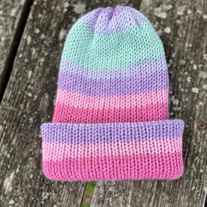 May include: A knitted beanie hat in a color block design. The hat features horizontal stripes of light green, lavender, pink, and fuchsia. The brim is folded up, and the hat is made of a soft, textured yarn.