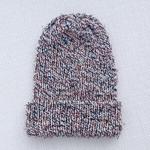 May include: A knitted beanie hat in a speckled pattern of white, brown, and black. The hat has a folded brim and a textured appearance. The hat is made of a soft, warm material, perfect for cold weather.
