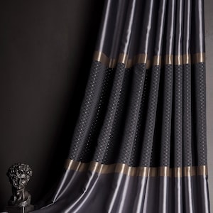 May include: Dark gray satin curtains with a textured, patterned section and gold trim. The curtains are draped, with soft folds and a lustrous sheen. A dark sculpture is visible in the background, suggesting a luxurious interior design theme.