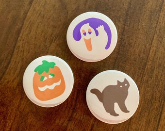 May include: Three round white buttons with Halloween themed illustrations. The first button has a purple ghost with a white face. The second button has an orange pumpkin with a green stem and a jack-o-lantern face. The third button has a black cat with white eyes.