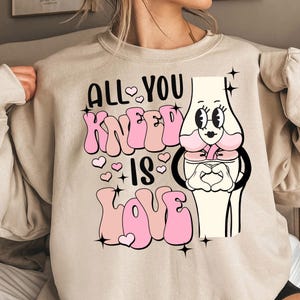 Puede incluir: Una sudadera beige con un dise&ntilde;o gr&aacute;fico rosa que dice "All You Kneel Is Love" con una ilustraci&oacute;n de rodilla de dibujos animados.