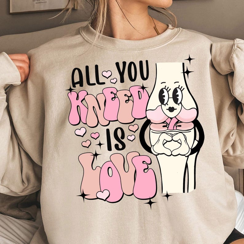 Puede incluir: Una sudadera beige con un dise&ntilde;o gr&aacute;fico rosa que dice "All You Kneel Is Love" con una ilustraci&oacute;n de rodilla de dibujos animados.