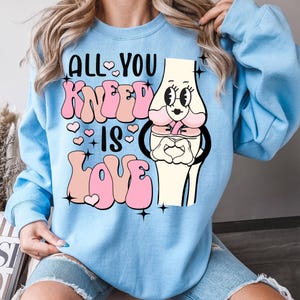Puede incluir: Sudadera azul claro con un hueso de rodilla de dibujos animados que lleva un lazo rosa y sostiene un coraz&oacute;n. El texto de la sudadera dice "All You Kneеd Is Love" en letras rosas y blancas.