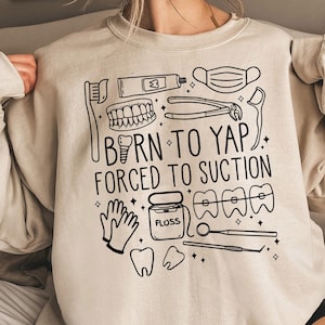 May include: Beige sweatshirt with a black graphic design featuring dental tools, teeth, and the text "BORN TO YAP, FORCED TO SUCTION." The design includes a toothbrush, toothpaste, and dental floss.
