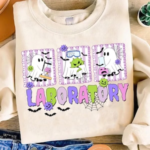 May include: Beige sweatshirt with a colorful "Laboratory" graphic. The graphic features cartoon ghosts, lab equipment, bats, and spiderwebs. The design has a Halloween or science theme.