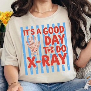 Puede incluir: Camiseta beige con el texto rojo "IT'S A GOOD DAY TO DO X-RAY", una mano esquelética y un símbolo de radiación. El diseño está sobre un fondo de rayas verticales azules.