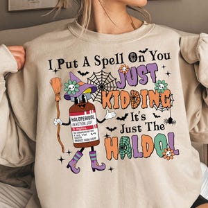 May include: A beige sweatshirt with a Halloween-themed graphic. The graphic features a cartoon witch holding a bottle labelled "HALOPERIDOL INJECTION, USP" and a broom. The text "I Put A Spell On You JUST KIDDING It's Just The HALDOL" is printed on the sweatshirt.
