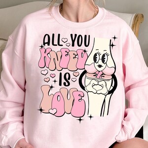 Puede incluir: Sudadera rosa con una ilustraci&oacute;n de rodilla de dibujos animados y el texto "All You Knead Is Love" en letras rosas y blancas con corazones y brillos.