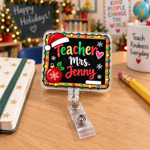May include: A square badge reel featuring a black chalkboard design with a colourful string of Christmas lights around the border. The text reads Teacher Mrs. Jenny in white and red script, accented by a Santa hat, a red ornament, and snowflakes.