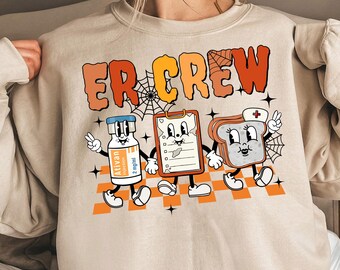 Könnte beinhalten: Ein beiger Pullover mit dem Text "ER CREW" in orangefarbenen und roten Buchstaben. Der Pullover zeigt eine Cartoon-Illustration einer Ampulle, einer Klemmbrett und einer Scheibe Brot mit einer Krankenschwestermütze, alle mit Gesichtern, Armen und Beinen.